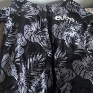 Svart vindjacka med vita blad från Svea  - Säljer en svart vindjacka från Svea  med vita tropiska bladmönster. Jackan har dragkedja framtill och huva. Perfekt för blåsiga dagar och enkel att packa ner i väskan.