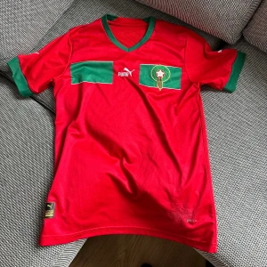 Marocko borta tröja 2022  - Marockos landslagströja för hemmamatcher under 2022. Klassisk röd färg med gröna detaljer vid ärmar och krage. Puma-logga på bröstet samt landslagets emblem. Tunt och andningsbart material som gör den skön att bära i alla väder. Ett måste för fans av Marockos landslag