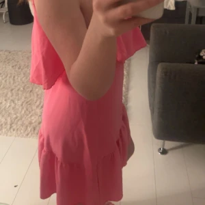 Rosa volangklänning med smala axelband - Säljer en söt rosa klänning med volangdetalj upptill och smala axelband. Klänningen har en avslappnad passform och är kort i modellen, perfekt för varma dagar.