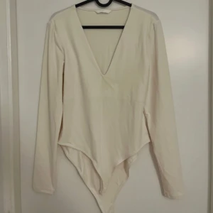 Vit långärmad bodysuit med v-ringning från H&M - Säljer en stilren vit bodysuit från H&M med lång ärm och djup v-ringning. Perfekt att styla till olika outfits. Mjukt och stretchigt material för en bekväm passform. Den är lite offwhite 