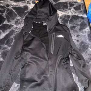 Svart softshelljacka med huva från The North Face - Säljer en svart softshelljacka från The North Face med huva och dragkedja. Jackan har en sportig look, diskret logga på bröstet och en bröstficka med dragkedja. Perfekt för utomhusaktiviteter och vardagsbruk.