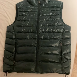 Mörkgrön dunväst från Polo Ralph Lauren - Snygg mörkgrön dunväst från Polo Ralph Lauren med dragkedja framtill och två sidofickor med dragkedja. Klassisk broderad logga på bröstet och justerbar dragsko nedtill. Perfekt för lager-på-lager under kyliga dagar.