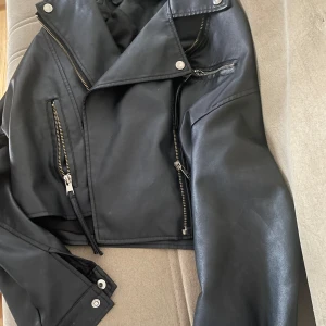 Svart skinnjacka med dragkedjor - Säljer en svart skinnjacka med coola detaljer som flera dragkedjor och tryckknappar. Jackan har en klassisk bikerstil med bred krage och croppad passform. Perfekt för dig som vill ha en edgy look. den är lite kortare i modellen och ena fickan har ett hål inuti, inget som syns på utsidan. passar en xs/s. står ingen lapp inuti