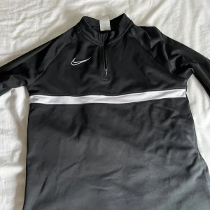 Svart träningströja från Nike - Säljer en svart långärmad träningströja från Nike med vit logga och en vit rand över bröstet. Tröjan har dragkedja vid halsen och är tillverkad i ett syntetmaterial som andas. Perfekt för träning eller sportaktiviteter.