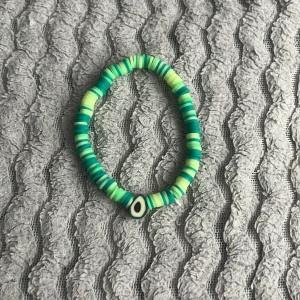 Grönt pärlarmband med evil eye detalj - Säljer ett färgglatt armband med gröna och turkosa platta pärlor samt en evil eye-pärla i mitten. Perfekt accessoar för att liva upp din outfit!