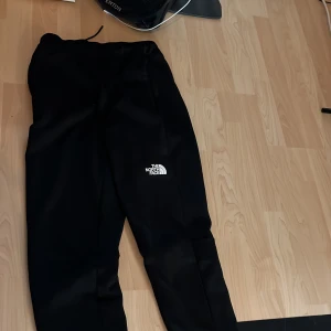 Svarta mjukisbyxor från The North Face - Svarta mjukisbyxor från The North Face med vit logga på låret. Byxorna har elastisk midja och är i en bekväm, avslappnad passform. Perfekta för chill eller träning.