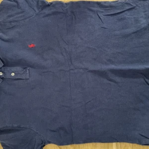 Blå pikétröja från Ralph Lauren - Mörkblå pikétröja från Ralph Lauren med klassisk krage och två knappar framtill. Liten röd broderad logga på bröstet. Perfekt för en avslappnad men ändå stilren look.