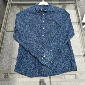 Blå blommig skjorta från GANT - Stilren och klassisk Gant-skjorta i bra skick. Perfekt för både vardag och lite finare tillfällen. Den här skjortan ger en sofistikerad look med sin slimmade passform och kvalitativa material. Ett bra komplement till garderoben för den modeintresserade!   Skick: Mycket bra. Passform: Normal. Storlek: M
