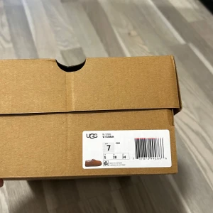 Bruna UGG tofflor med mönstrad kant - Säljer ett par bruna UGG tofflor i mocka med mjuk ullfodring. Skorna har en dekorativ mönstrad kant i svartvitt och en rejäl, mönstrad sula för bra grepp. Perfekta för att hålla fötterna varma inomhus eller utomhus. 