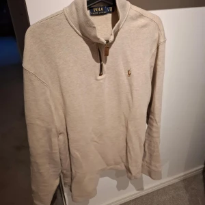 Beige halvzip tröja från Polo Ralph Lauren - Säljer en stilren beige långärmad tröja från Polo Ralph Lauren med halv dragkedja och broderad logga på bröstet. Perfekt för en avslappnad men snygg look. Hög krage och mjukt material.