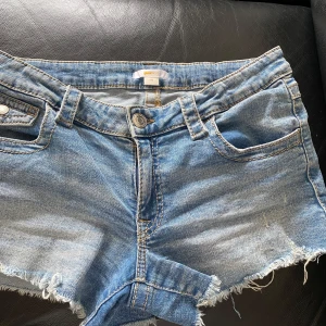 Blå jeansshorts, lågmidjade ,  från Gina Tricot - Säljer ett par klassiska lågmidjade blå jeansshorts från Gina Tricot. Shortsen har fransade benslut, fem fickor och knappgylf. Perfekta för sommaren och har en snygg, lite sliten look., stl 170. Mätt rakt över 37-38 cm. Går att spänna in! 