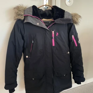 Svart vinterjacka med fuskpäls från Peak Performance - Säljer en svart vinterjacka från Peak Performance med huva och fuskpäls. Jackan har rosa dragkedjor, flera fickor och patch på ärmen. Perfekt för kalla dagar och snygg detalj med pälsen på huvan.