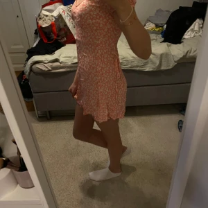 Rosa blommig klänning från Cotton On - Söt kortklänning från Cotton On i ljusrosa med vitt blommönster. Klänningen har kort ärm, v-ringning och volangdetalj nertill. Perfekt för varma dagar och har en feminin, lätt och luftig känsla.