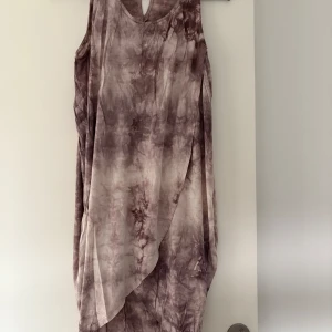 Lång batikmönstrad klänning  - Säljer en lång, ärmlös klänning från Gina Tricot i batikmönster med lila och beige toner. Klänningen har en avslappnad passform och v-ringning framtill. Perfekt för varma dagar och enkel att styla med accessoarer.