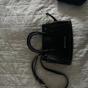 Svart handväska från Valentino - Snygg svart handväska från Valentino med både handtag och justerbar axelrem. Väskan har guldfärgade detaljer och stilren design med logga på framsidan. Perfekt för att bära dina viktigaste saker med stil.