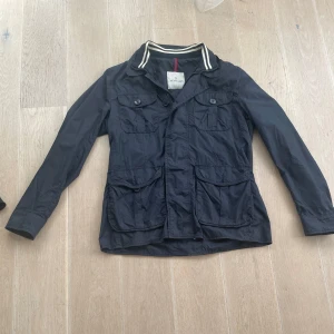 Moncler jacka  - Moncler-jacka i marinblått – lätt, stilren och perfekt för vår och höst. Klassisk modell med fyra fickor och sportig krage. Använd få gånger, i riktigt fint skick. Säljer för att den inte kommer till användning längre.