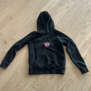 Svart hoodie från GANT med logga - Svart hoodie från GANT med broderad logga och sköld på bröstet. Klassisk huva och magficka framtill. Perfekt för en avslappnad stil och enkel att matcha med det mesta.