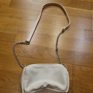 Vit axelväska med kedja - Säljer en stilren vit axelväska med silverfärgad kedja och dragkedja upptill. Väskan har en mjuk, slät yta och en enkel, modern design som passar till många olika outfits.
