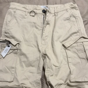 Beige cargoshorts i linne från Mouli - Säljer ett par beige cargoshorts i linne från Mouli med flera fickor, dragkedja och knapp framtill. Shortsen har bälteshällor och är tillverkade i bomull med lite stretch för extra komfort. Perfekta för sommardagar när du vill ha en avslappnad stil.