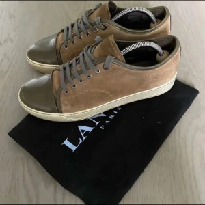Bruna sneakers från Lanvin med grå detaljer - Snygga sneakers från Lanvin i brunt mocka och grått skinn. Skorna har grå snörning, rund tå med glansig grå tåhätta och vit platt sula. Perfekta för dig som vill ha en stilren och exklusiv look.