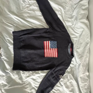 Mörkblå stickad tröja med amerikansk flagga från GANT - Säljer en mörkblå stickad tröja från GANT med en stor amerikansk flagga på bröstet och broderad text på ärmen. Tröjan har ribbade muddar och rund hals. Perfekt för dig som gillar klassisk preppy-stil.