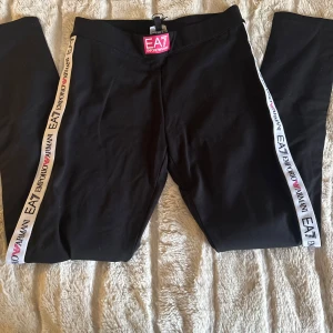 Svarta leggings från EA7 Emporio Armani - Svarta leggings med vita sidor där det står 'EA7 Emporio Armani' och små rosa hjärtan. Bekväm passform och elastiskt material, perfekt för en sportig look. Midjan är normal och loggan syns tydligt både på sidan och i midjan.