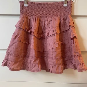 Rosa volangkjol från ONLY  - Supersöt rosa kjol från ONLY med volanger och smock i midjan. Kjolen har fina spetsdetaljer och är tillverkad i ett lätt och luftigt tyg, perfekt för varmare dagar. Passar dig som gillar romantisk och feminin stil. Kjolen ör oanvänd och säljs då jag inte trivs i färgen 