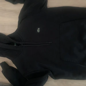 Svart hoodie från Lacoste - Säljer en svart hoodie från Lacoste med klassisk krokodillogga på bröstet. Tröjan har huva med dragsko och en stor magficka framtill. Perfekt för en avslappnad och stilren look.