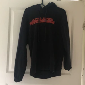 Svart hoodie från Jack & Jones - Svart hoodie från Jack & Jones med röd logga på bröstet. Tröjan har långa ärmar, huva med dragsko och är tillverkad i mjukt material. Perfekt för en avslappnad stil.