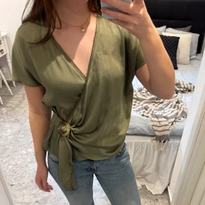 Omlottopp med ringdetalj - Omlottopp från Zara Basic i glansigt material. Toppen har korta ärmar, v-ringning och en dekorativ ring i midjan där tyget knyts. Mycket bra skick!