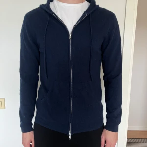 Mörkblå hoodie i kashmir från John Henric - Säljer en mörkblå hoodie i mjuk kashmir från John Henric. Tröjan har dragkedja framtill, huva med dragsko och långa ärmar. Perfekt för dig som gillar stilrena och bekväma plagg.