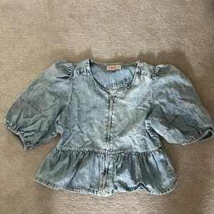 Ljusblå jeansblus med puffärm från ONLY - Supersöt ljusblå jeansblus från ONLY med puffiga korta ärmar och volang nertill. Blusen har rund halsringning och dragkedja framtill. Perfekt för dig som gillar denim och vill ha en trendig topp med lite extra detaljer.