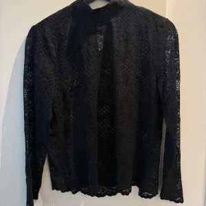 Svart spetsblus med lång ärm - Säljer en svart blus i genombruten spets med lång ärm. Blusen är transparent och har ett elegant mönster i tyget. Perfekt att styla lager på lager eller med ett linne under.