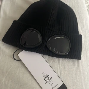 Svart mössa med glasögondetalj från C.P. Company - Svart ribbstickad mössa från C.P. Company med unika glasögondetaljer framtill. Märkets logga syns på ena linsen. Perfekt accessoar för en cool och modern look.