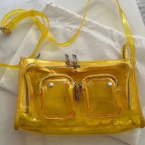 Gul transparent axelväska från Núnoo - Gul transparent axelbandsväska från Núnoo med dragkedja, justerbar rem och flera fack framtill samt ett fack inuti. Säljer då den inte kommit till användning. Bra skick/ använd en gång. Köpt för 1299kr på Åhléns Perfekt nu till våren och nu när gult är årets sommarfärg!!💛