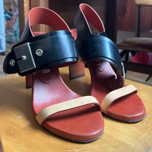 Unika klackade sandaler från Acne Studios - Säljer ett par statement sandaler från Acne Studios med bred svart rem och stor silverdetalj. Skorna har rödbrun sula, beige rem över tårna och hög klack. Perfekta för dig som gillar en edgy och modern stil.