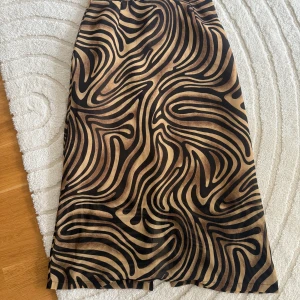 Zebramönstrad långkjol med knappdetalj - Säljer en snygg långkjol med zebramönster i svart och beige. Kjolen har en knappdetalj i midjan och är tillverkad i ett lätt, glansigt material som ger ett coolt intryck. Perfekt för dig som vill sticka ut med ett djurmönster.