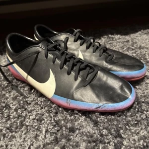 Svarta Nike fotbollsskor med vit swoosh - Säljer ett par svarta Nike fotbollsskor med stor vit swoosh på sidan. Skorna har snörning och en platt sula med dobbar i svart och rosa undertill. Perfekta för dig som vill ha stilrena och sportiga skor på planen.