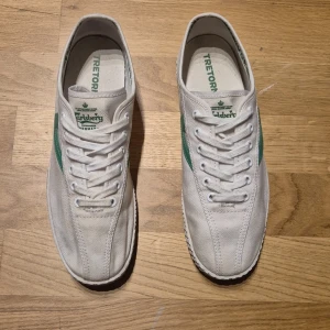 Vita Tretorn x Carlsberg sneakers med gröna detaljer - Säljer ett par vita Tretorn sneakers i samarbete med Carlsberg. Skorna har gröna detaljer på sidan och loggor på plösen och innersulan. Klassisk låg modell med snörning och platt sula. Perfekta för dig som gillar retrostil och unika samarbeten.