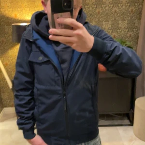 Mörkblå vindjacka med dragkedja - Säljer en mörkblå vindjacka med huva och dragkedja framtill. Jackan har en bröstficka med knapp och två sidofickor. Perfekt för blåsiga dagar och enkel att matcha med olika outfits.