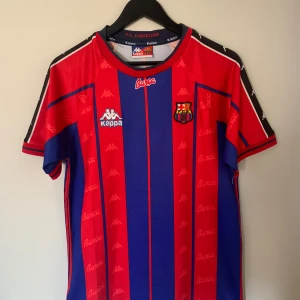 FC Barcelona fotbollströja från Kappa - Klassisk FC Barcelona fotbollströja från Kappa med röda och blå vertikala ränder, klubbmärke på bröstet och Kappa-loggor på axlarna. Kortärmad modell med rund hals och tryckt 'Barça' på framsidan. Tröjan är ifrån 1997-98 säsongen.