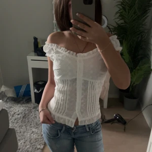 Vit spetsblus med volang - Säljer en söt vit blus med spetsdetaljer och volang vid axlarna. Blusen är axelbandslös och har knappar framtill. Perfekt till jeans eller kjol för en fräsch look.