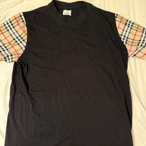 Burberry sommarset - Svart t-shirt med klassiska beige, röda och svarta rutiga ärmar. T-shirten har rund halsringning och normal passform. Perfekt för dig som vill ha en stilren look med en twist.