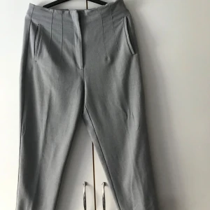 Grå byxa med hög midja - Snygga grå kostymbyxor med hög midja och raka ben. Byxorna har diskreta fickor framtill och pressveck som ger en stilren look. Perfekta till en dressad outfit.