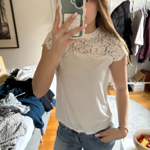 Vit topp med spetsdetaljer - Säljer en söt vit topp med spets över axlar och bröst. Toppen har korta ärmar och en rund halsringning. Perfekt för dig som gillar romantiska detaljer och en feminin stil.