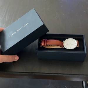 Klassisk klocka från Daniel Wellington med brunt läderband - Stilren klocka från Daniel Wellington med vitt ur och brunt läderarmband. Urtavlan är rund och har silverfärgade detaljer. Perfekt accessoar för en clean och tidlös look. Den är helt ny, en 50års present. Kan gå ner i pris vid snabb affär🙌 nypris 1999kr