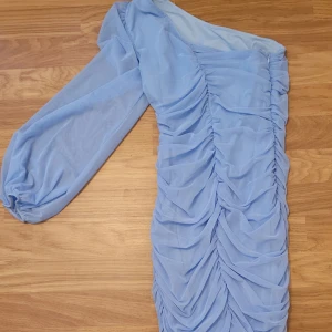 Ljusblå one shoulder klänning med rynk - Trendig ljusblå klänning med one shoulder-design och långa puffärmar. Klänningen har snygga rynkdetaljer framtill och baktill som ger en cool och modern look. Perfekt för dig som vill sticka ut med en unik stil.