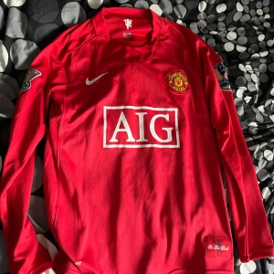 Manchester United långärmad fotbollströja 07/08 - Klassisk röd Manchester United långärmad matchtröja från säsongen 07/08 med AIG som sponsor, broderad klubbmärke och Premier League-märke på ärmen. Materialet är NikeFIT för bra ventilation. 'The Red Devils' detalj nertill.