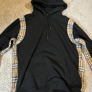 Svart hoodie med rutiga detaljer - Säljer en svart hoodie med klassiska Burberry-rutiga paneler längs ärmarna och sidorna. Tröjan har huva  och en stor magficka framtill. Perfekt för en trendig och avslappnad stil. Skriv vid minsta fundering och pris går att diskuteras 
