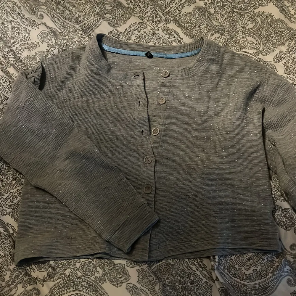 Säljer en grå croppad kofta med diskret glittrigt material och knappar framtill. Koftan har långa ärmar och en enkel, stilren design. Perfekt att slänga på sig över en topp för en extra touch av glitter. Koftan har relativt korta ärmar.. Neuletakit & Villapaidat.